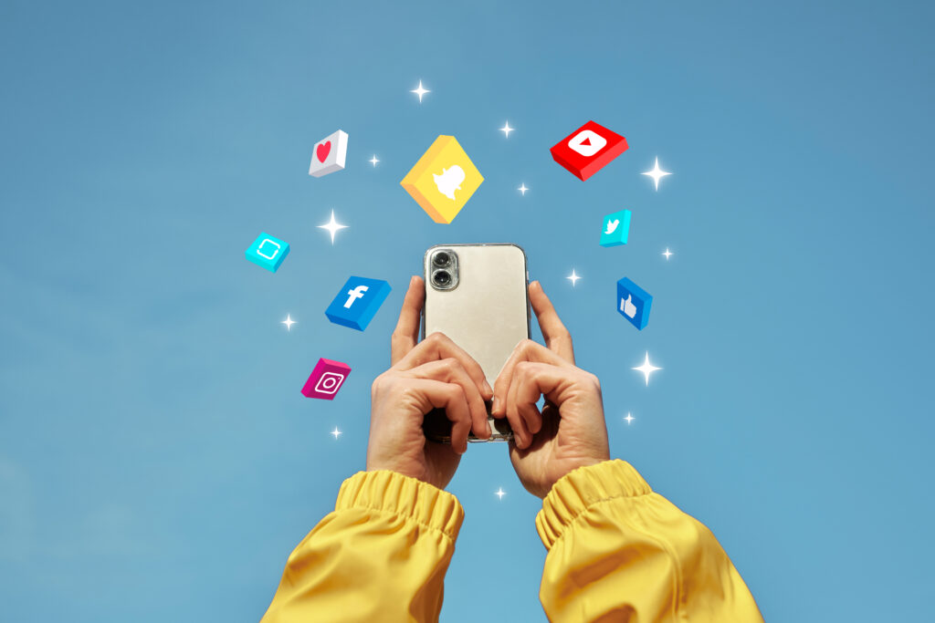 Das Bild zeigt zwei Hände in gelben Ärmeln, die ein silberfarbenes Smartphone in die Luft heben. Der Hintergrund ist blau und um das Smartphone herum schweben animierte Social Media Icons.